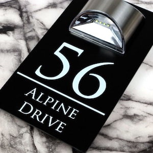 May include: Black acrylic house number sign with white lettering. The sign reads "56 Alpine Drive". A small, silver, solar-powered light is mounted above the house number.