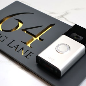 May include: A black address sign with the number 64 cut out, revealing a gold backing. The words "ING LANE" are below the numbers. A silver Ring doorbell is next to the sign.