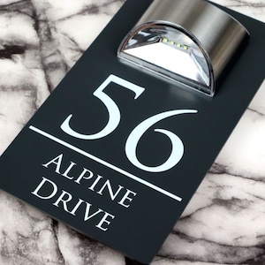 以下が含まれることがあります： 白い文字で書かれた黒い家の番号札。札には「56 Alpine Drive」と書いてあります。家の番号の上に小さな銀色のソーラーライトが取り付けられています。