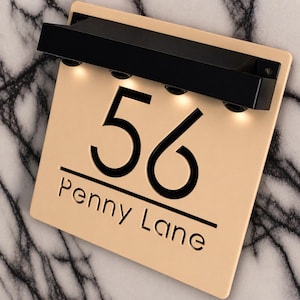 Può includere: Una targa quadrata color beige per numero civico con il numero 56 e il testo Penny Lane incisi in nero. La targa include una lampada orizzontale nera montata sulla parte superiore con quattro luci LED integrate rivolte verso il basso.
