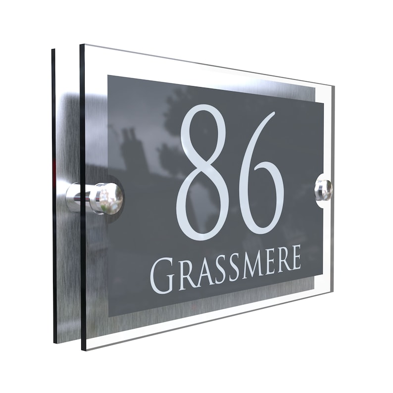 Puede incluir: Un letrero de n&uacute;mero de casa de acr&iacute;lico transparente y plateado con el n&uacute;mero 86 y el nombre Grassmere en letras blancas sobre un fondo gris.