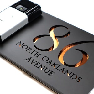 Könnte beinhalten: Schwarzes Hausnummernschild mit der ausgeschnittenen Nummer 86, die von hinten beleuchtet wird. Das Schild trägt außerdem die Aufschrift "North Oaklands Avenue".