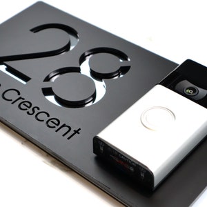 Puede incluir: Un letrero de número de casa negro con el número 28 y la palabra "Crescent" grabada en él. Una cámara de timbre plateada está montada debajo del letrero.