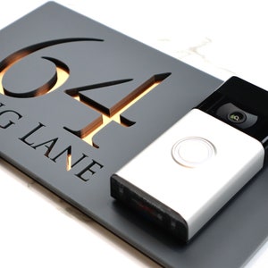 May include: A black and gold house number sign with the number 64 and the text "King Lane" on a gray background. A silver doorbell camera is mounted below the sign.