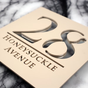 Può includere: Un cartello di numero civico in legno beige con il numero 23 ritagliato e le parole "Honeysuckle Avenue" sotto.