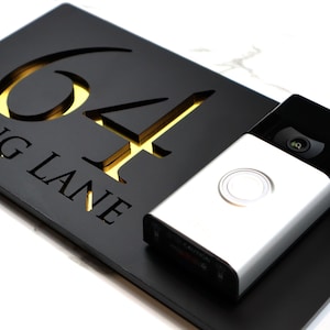 May include: Black house number sign with gold numbers "64" and the text "ING LANE" engraved on the surface. A silver doorbell camera is mounted on the sign.