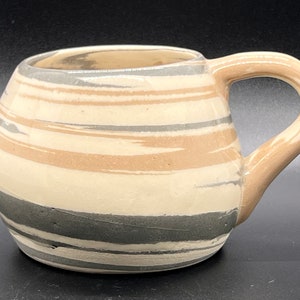 May include: A ceramic mug with a rounded body and a handle. The mug is decorated with a striped pattern in shades of gray, brown, and white.