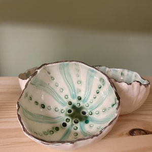 May include: Three handmade ceramic bowls. The bowls are white with a teal and green pattern inside, and a dark grey rim. The bowls are arranged on a light wood surface. Each bowl has a unique, organic shape.