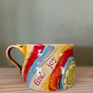 May include: A ceramic mug with a vibrant, abstract design. The mug has a yellow base with red, blue, and orange stripes. The words "find joy" are written in red. The handle is yellow with a brown accent.