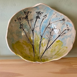 May include: A handcrafted ceramic bowl with an irregular shape. The interior is decorated with a painted design of black floral stems set against a backdrop of blue and green colours, creating a natural look. The bowl's rim is a creamy white.