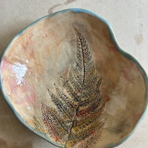 May include: Handmade ceramic bowl with a fern design. The bowl has a light brown interior with a detailed fern illustration in black, red, and yellow. The rim is outlined in a light blue colour.