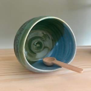 May include: A handcrafted ceramic bowl with a blue and green glaze, on a wooden surface. A small wooden spoon is inside the bowl. The bowl's interior has a swirling pattern.