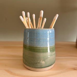 May include: A small, cylindrical ceramic vessel with a glossy finish, featuring a horizontal colour gradient of light blue at the top and sage green at the bottom. The vessel is filled with several wooden matches that have white tips.