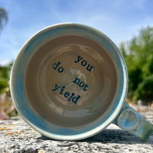 May include: A ceramic mug with a light blue glazed rim and a cream-coloured interior. The words "you do not yield" are printed in dark blue lettering on the bottom inside of the mug. The mug features a sturdy handle on the right side.