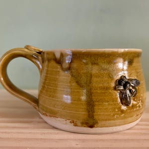 May include: A handmade ceramic mug with a yellow-brown glaze and a white base. The mug features a sculpted bee on the side and a curved handle. The mug is suitable for hot or cold beverages.