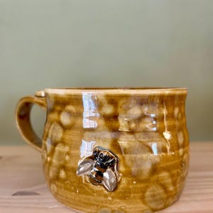May include: A round ceramic mug with a glossy, mottled honey-brown glaze featuring a textured surface. A small, three-dimensional sculpted bee with black and yellow stripes and silver-toned wings is attached to the front of the mug body.