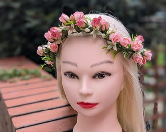 May include: A pink rose flower crown with green leaves and white accents. The floral crown is displayed on a mannequin head with blonde hair and red lipstick. The background is blurred.
