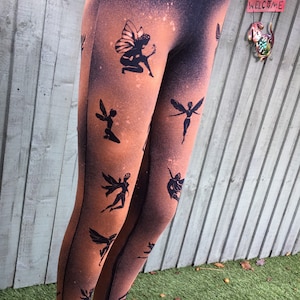 May include: Black leggings with a faded orange and brown splatter pattern. The leggings feature a repeating pattern of black fairy silhouettes.