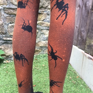 May include: A pair of brown tights with a black spider design. The tights are ombre, fading from a darker brown at the top to a lighter brown at the bottom.