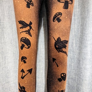 May include: Black leggings with a brown, bleached effect. The leggings are patterned with black silhouettes of mushrooms, fairies, and arrows. The design is repeated down the legs, creating a whimsical pattern.