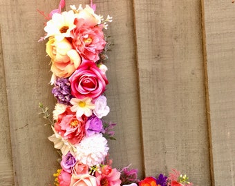 May include: A pink, white, and purple floral letter L made of artificial flowers. The letter is hanging from a pink ribbon with green leaves.