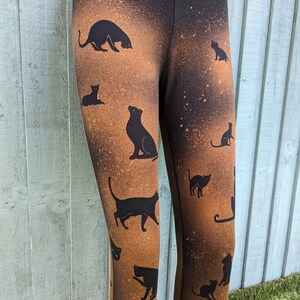 May include: Black leggings with a brown, speckled design and black cat silhouettes. The cats are in different poses, such as sitting and standing. The leggings are made of a soft, stretchy fabric.