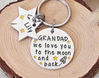 May include: A silver-toned metal keychain featuring a circular disc engraved with the text "GRANDAD we love you to the moon and back" and a small rocket ship, accompanied by two star-shaped charms, one of which is engraved with the name "Lily".
