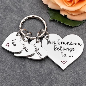 May include: A silver-toned metal keyring featuring a large heart-shaped charm engraved with the text This Grandma Belongs To... and three smaller heart-shaped charms engraved with the names Lea, Ava, and Joshua, each accented with a small red heart.
