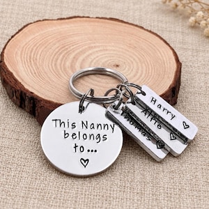 Pode incluir: Um porta-chaves de metal prateado com um disco circular grande gravado com o texto 'This Nanny belongs to...' e um símbolo de coração, acompanhado por três etiquetas retangulares menores gravadas com os nomes Harry, Alfie e Thomas.