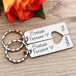 May include: Two rectangular silver-toned metal keychains featuring a cutout heart shape. Each keychain is engraved with the text Besties Forever and a heart symbol. One keychain is engraved with the name Ria and the other with the name Calvin.