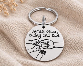 May include: A circular silver-toned metal keychain featuring an engraved design of three human fists and one animal paw in a fist-bump formation. The text James, Oscar, Buddy and Dad is engraved above the graphic illustration on the metal disc.