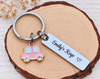 May include: A silver-toned metal keychain featuring a circular ring, a small pink car charm with light blue windows and clear crystal wheels, and a rectangular silver tag engraved with the text Emily's Keys followed by a small heart symbol.