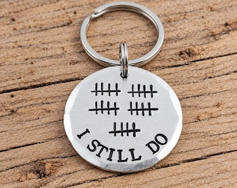 May include: A circular silver-toned metal keychain featuring a split ring at the top. The surface is engraved with five sets of tally marks arranged in a pattern above the black text that reads I STILL DO in a bold, sans-serif capital font.