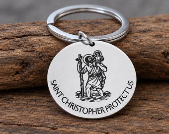 May include: A circular silver-toned metal keychain featuring an engraved illustration of Saint Christopher carrying a child across water, with the text SAINT CHRISTOPHER PROTECT US inscribed along the bottom edge of the disc.