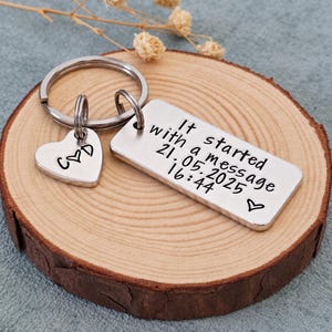 May include: A silver-toned metal keyring featuring a rectangular tag engraved with the text It started with a message 21.05.2025 16:44 and a small heart symbol, alongside a smaller heart-shaped charm engraved with the letters C and D with a heart.