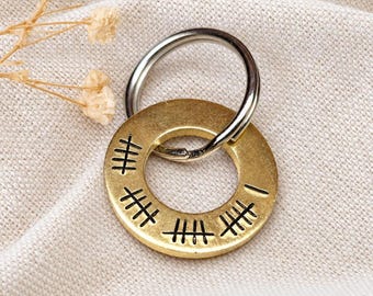 May include: A circular brass pendant featuring hand-stamped black tally marks around its surface, attached to a silver-toned metal split ring. The metal disc has a brushed finish and displays multiple sets of etched tally marks.