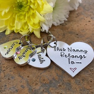 May include: Silver heart-shaped keychain with the text "This Nanny Belongs To..." and a small heart. Three smaller silver heart-shaped charms with the names "Carson", "Emily", and "Faith" are attached to the keychain.