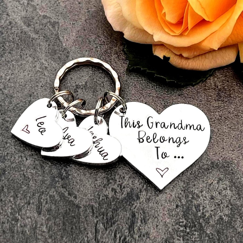 Pode incluir: Um porta-chaves de metal prateado com um pendente grande em forma de cora&ccedil;&atilde;o gravado com o texto "This Grandma Belongs To..." e um pequeno &iacute;cone de cora&ccedil;&atilde;o, acompanhado por tr&ecirc;s pendentes menores em forma de cora&ccedil;&atilde;o com os nomes "Leo", "Ava" e "Joshua".