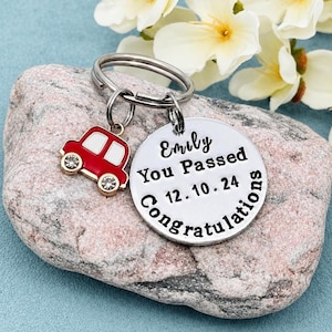 May include: A silver-toned metal keyring featuring a round disc engraved with the text Emily You Passed 12.10.24 Congratulations and a small red and white car charm with two clear rhinestones serving as wheels attached to the split ring.