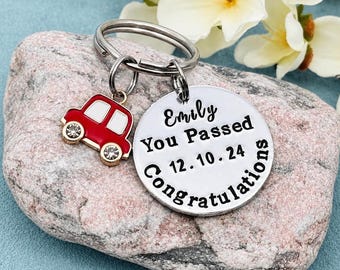 May include: A silver-toned metal keyring featuring a round disc engraved with the text Emily You Passed 12.10.24 Congratulations and a small red and white car charm with two clear rhinestones serving as wheels attached to the split ring.