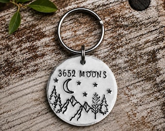 May include: A round, hammered silver-tone metal keychain featuring a stamped design of mountains, pine trees, stars, and a crescent moon. The text 3652 MOONS is stamped across the top. The keychain includes a circular metal split ring for attachment.