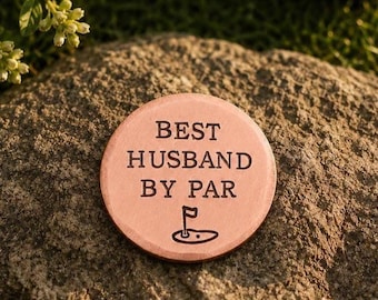 Puede incluir: Un marcador de pelota de golf redondo de metal color cobre con el texto negro grabado BEST HUSBAND BY PAR y un pequeño gráfico de una bandera de golf en un green, apoyado sobre una superficie de piedra texturizada e irregular.