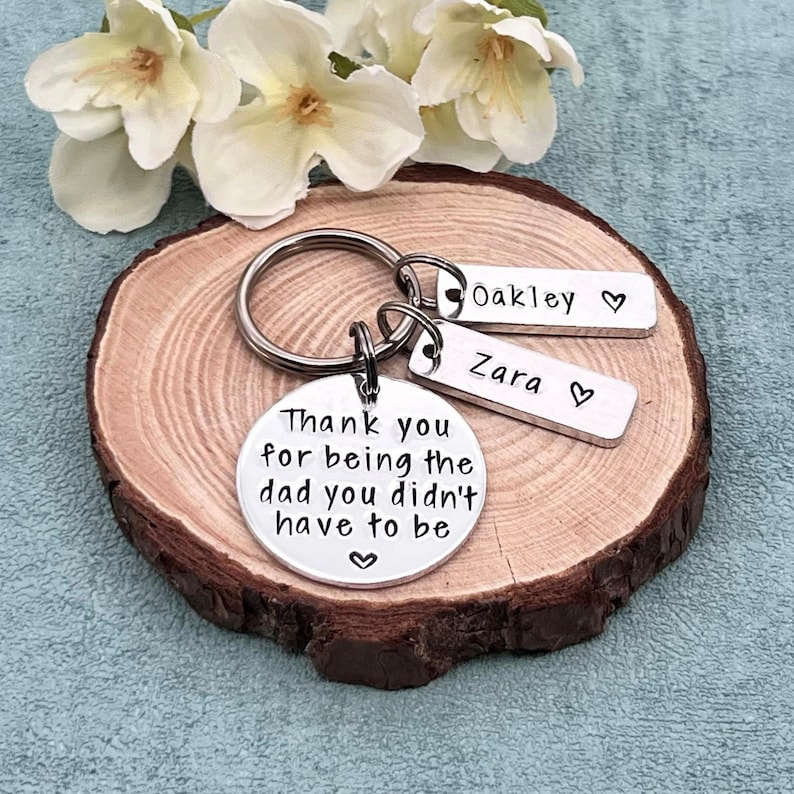 May include: Silver keychain with a round charm that reads "Thank you for being the dad you didn't have to be".  Two smaller charms with the names "Oakley" and "Zara" are attached to the keychain.