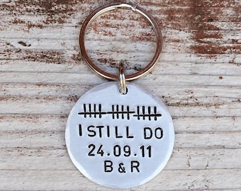 May include: A round silver-toned metal keychain featuring a split ring at the top. The disc is stamped with black text that reads I STILL DO, the date 24.09.11, the initials B & R, and a series of tally marks representing the number of years.