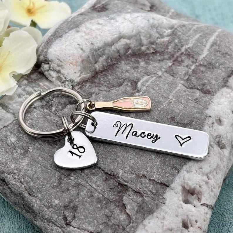 May include: Silver keyring with a heart charm that says "18", a champagne bottle charm, and a bar charm that says "Macey" with a heart.