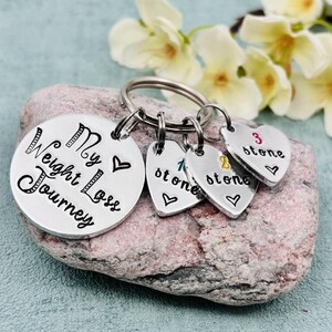 May include: A silver-toned metal keychain featuring a large round disc engraved with the text My Weight Loss Journey and a heart symbol. Three smaller heart-shaped charms are attached, each engraved with a number and the word stone.