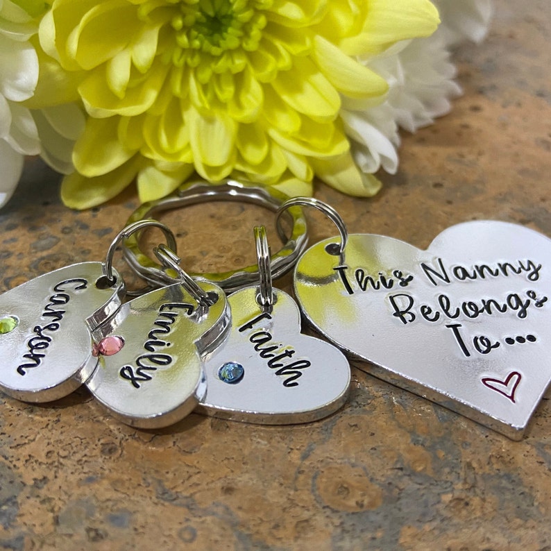 May include: Silver heart-shaped keychain with the text "This Nanny Belongs To..." and three smaller heart-shaped charms with the names "Carson", "Emily", and "Faith". Each charm has a small rhinestone.