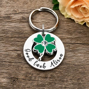 May include: A silver-toned circular keychain featuring a green four-leaf clover charm with a central clear crystal. The metal ring is engraved with the black text Good luck Alison, arranged in a curved script font along the bottom edge.