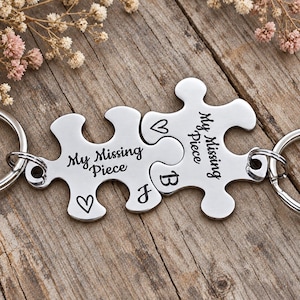Pode incluir: Dois porta-chaves de metal prateado em forma de peças de puzzle entrelaçadas. Cada peça está gravada com o texto My Missing Piece, um símbolo de coração e as letras J e B. Os porta-chaves possuem argolas metálicas circulares.
