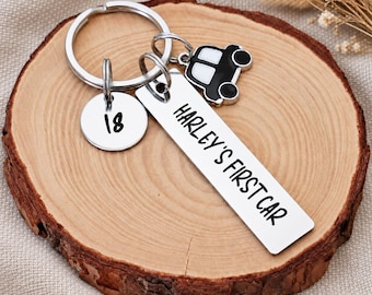 May include: A silver metal keychain featuring a round charm engraved with the number 18, a rectangular tag engraved with the text HARLEY'S FIRST CAR, and a small black and white enamel car charm attached to a circular split ring.